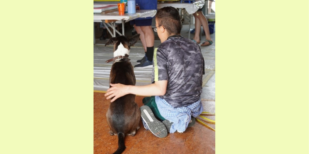 Mensch mit Behinderung und ein Hund sitzen kuschelnd am Boden. 