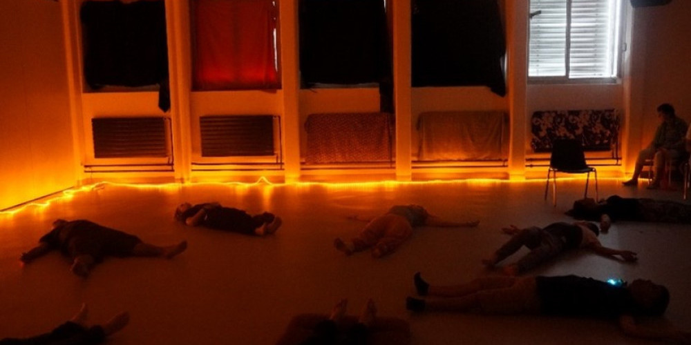 Menschen liegen in einem Keis angeordet am Boden. Eine Lichterkette beleuchtet den sonst dunklen Raum. 